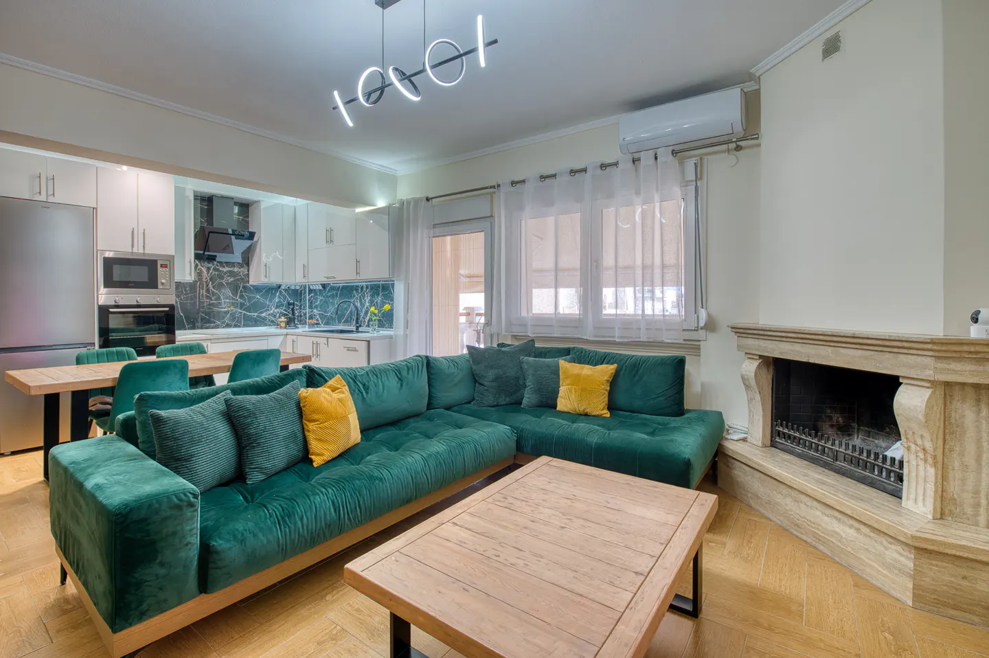 Bright living room with green sectional sofa, wood coffee table, fireplace, and open kitchen with white cabinets.