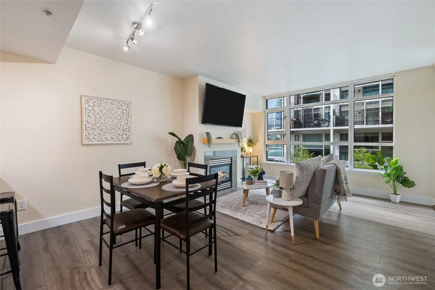 Living room with dining table, gray sofa, fireplace, and large window with city view. Wood floors and neutral walls.