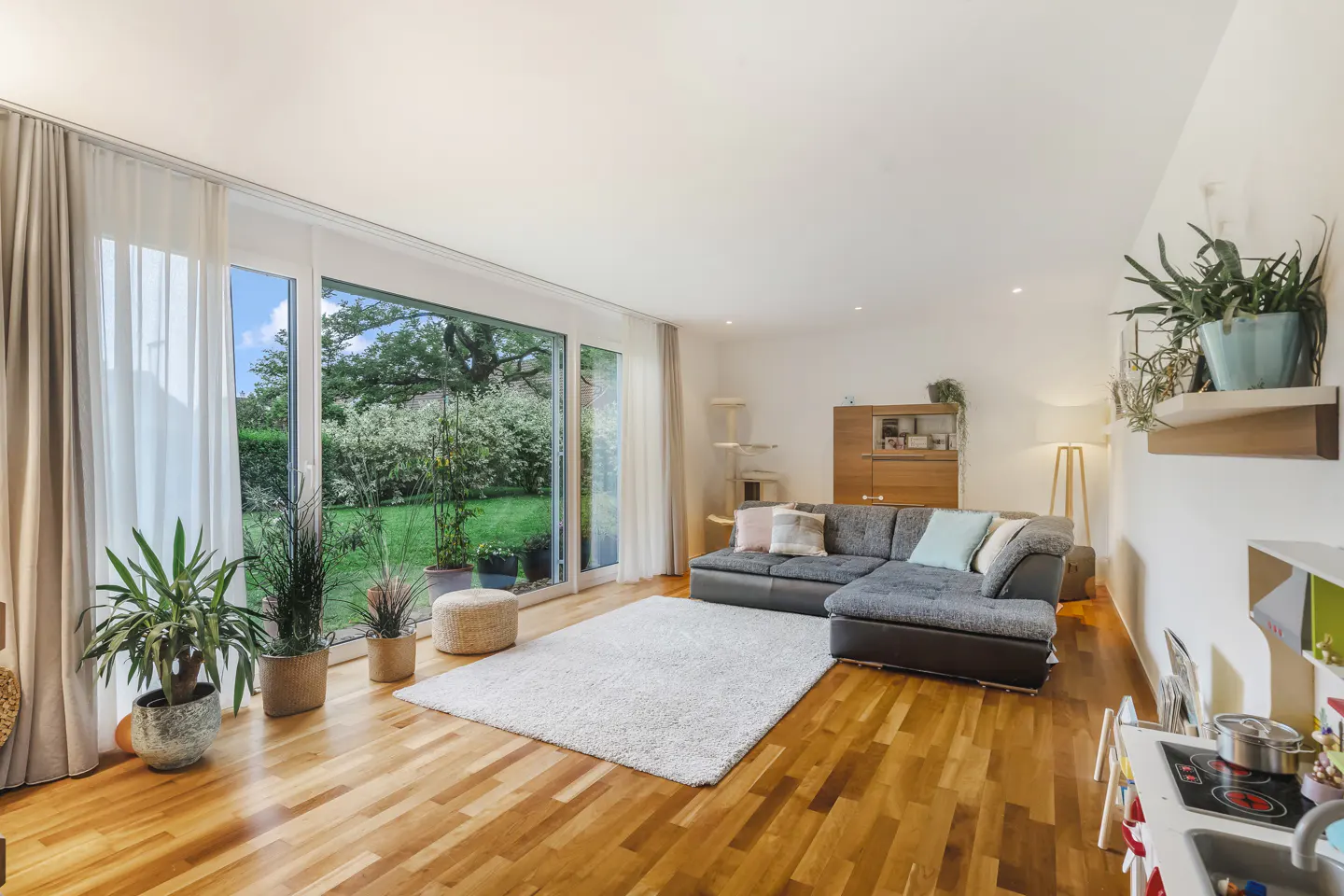 Bright living room with wood floors, a gray sectional sofa, and a large window overlooking a green garden. Plants add a touch of nature.