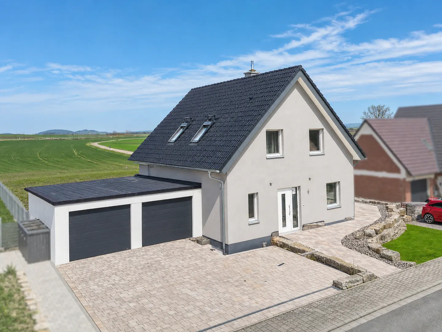 Two-story house with a gray exterior, black roof, and two-car garage on a paved driveway with a green field in the background.