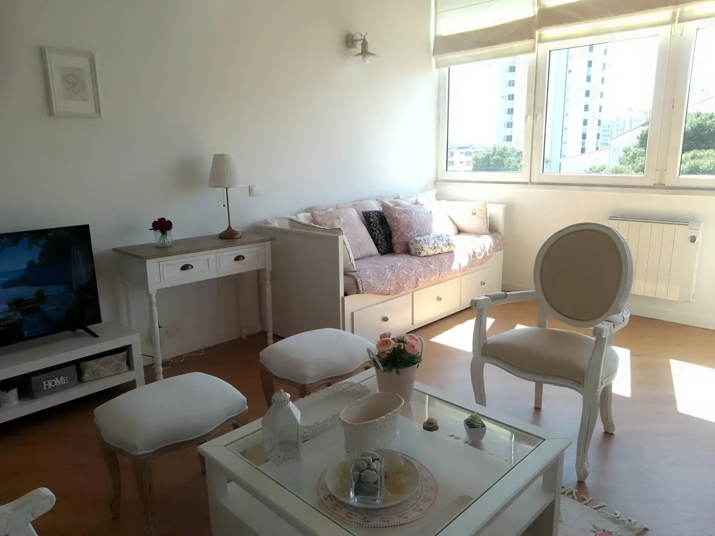 Bright living room with white furniture, including a daybed, chair, and glass-topped coffee table. Large windows offer a city view.