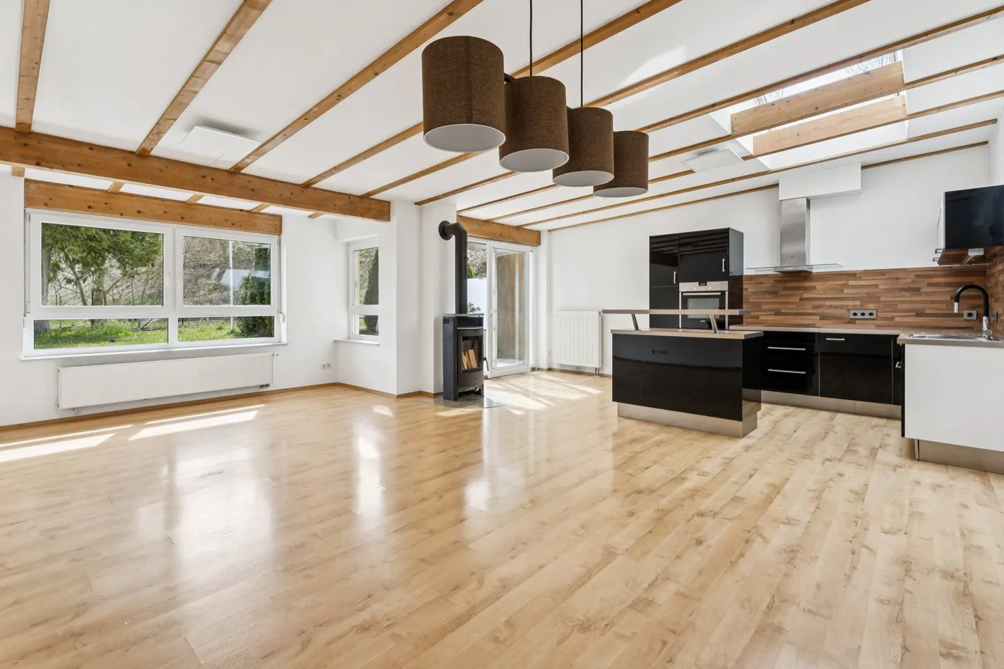 Bright, open-concept living space with wood floors, white walls, exposed beams, and a modern black kitchen. A wood-burning stove adds warmth.