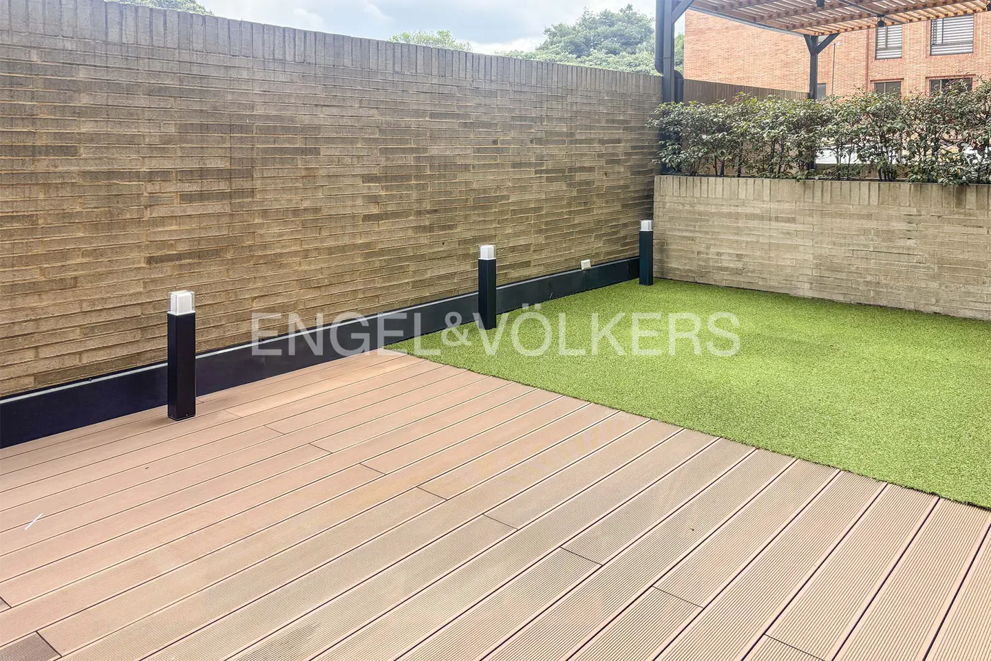 Outdoor patio with wood deck, green turf, and brick walls. Black light posts line the edge. Engel & Völkers logo is superimposed.