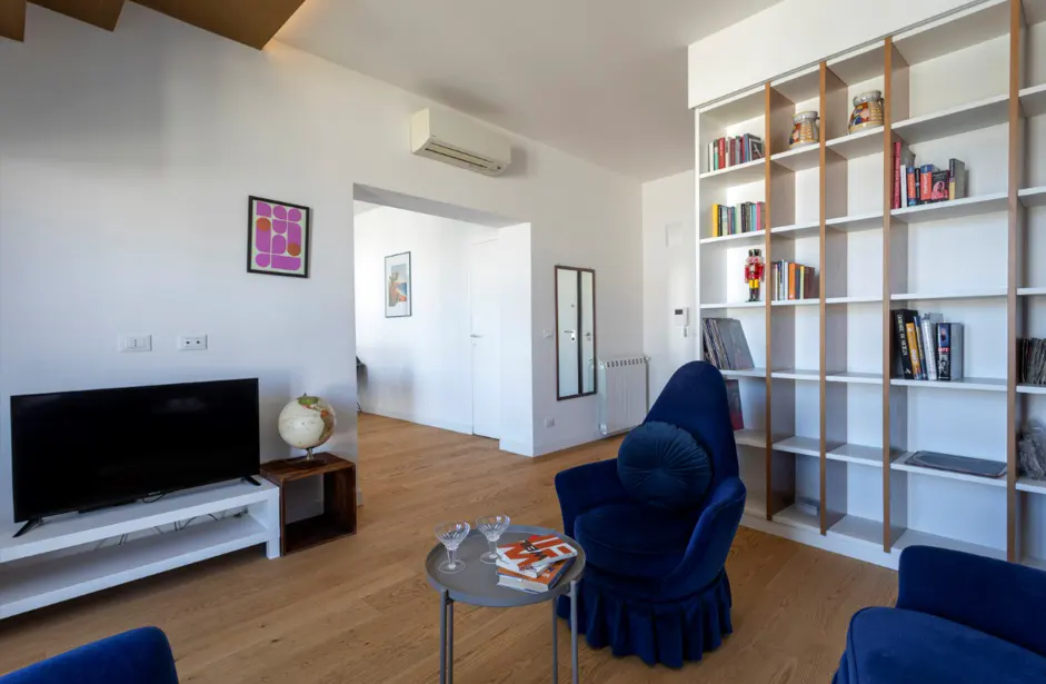Bright living room with wood floors, white walls, and blue velvet chairs. A bookshelf, TV, globe, and small table decorate the space.