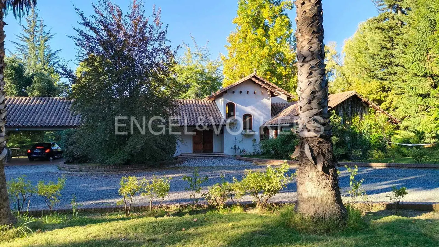 Exterior view of a light blue house with a brown tile roof, surrounded by trees and greenery on a sunny day.