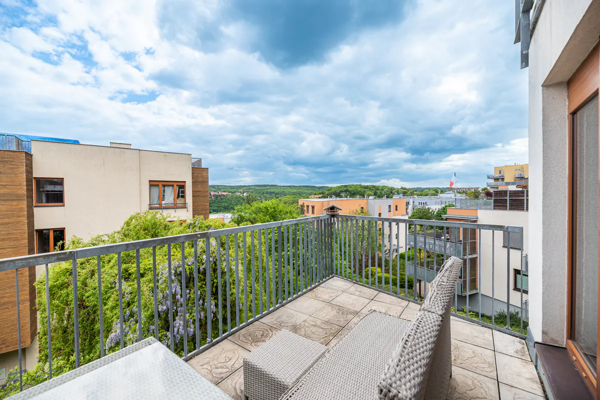 Balcony view with gray wicker furniture, metal railing, and tile floor. Lush greenery and buildings in the background under a cloudy sky.