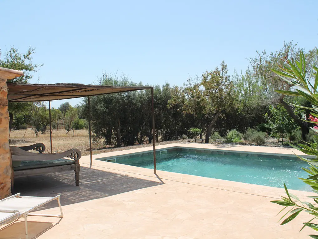 Outdoor pool with turquoise water, beige stone patio, and shaded seating area with a wooden bench and sun lounger. Trees and blue sky in the background.