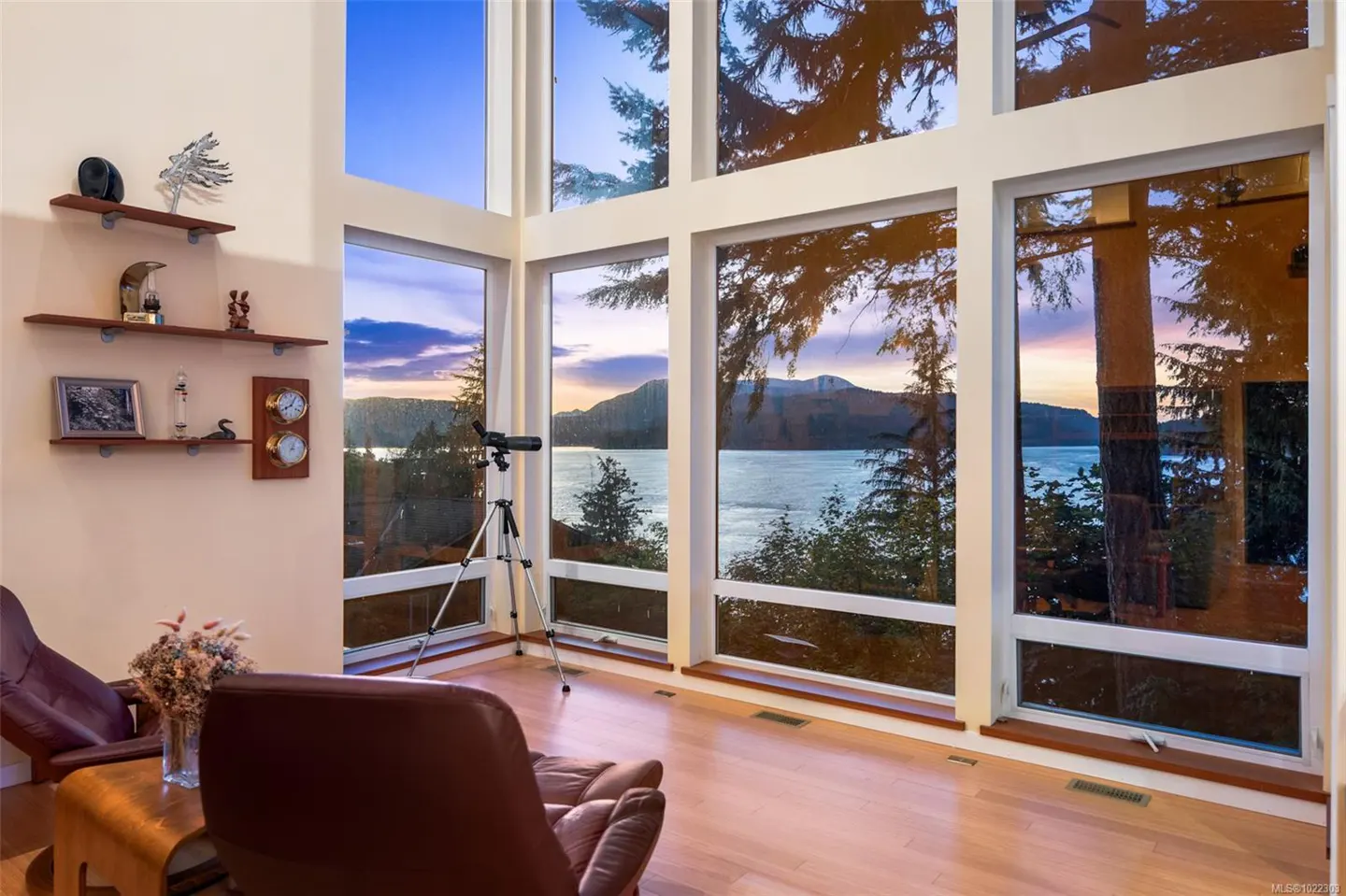 Living room with large windows overlooking a lake at sunset. Telescope on tripod, leather chairs, and shelves with decor.
