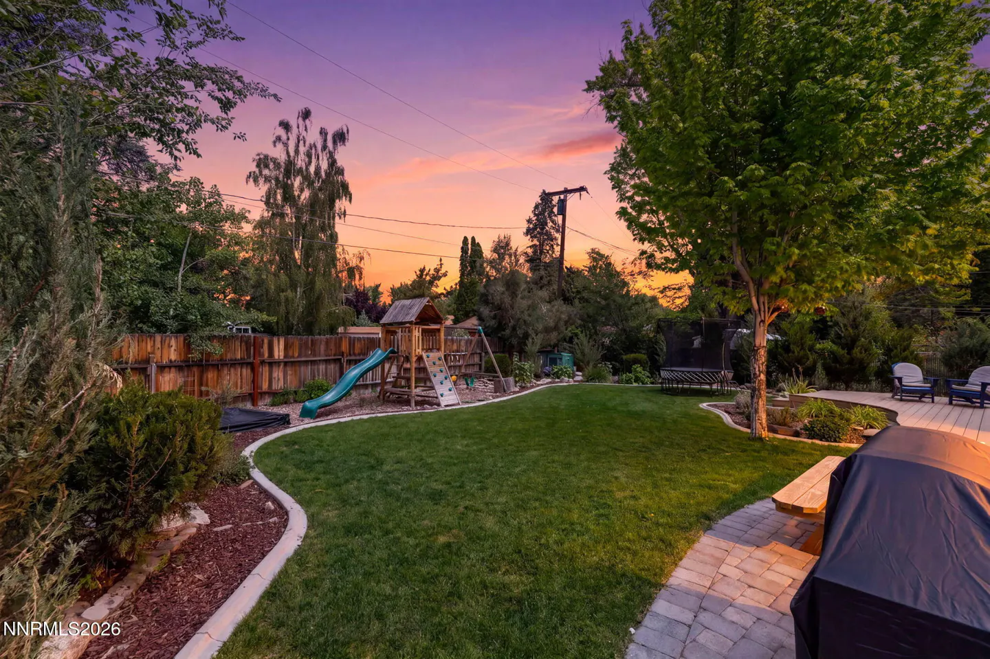 A backyard with green grass, a wooden fence, a playset, and a deck at sunset.
