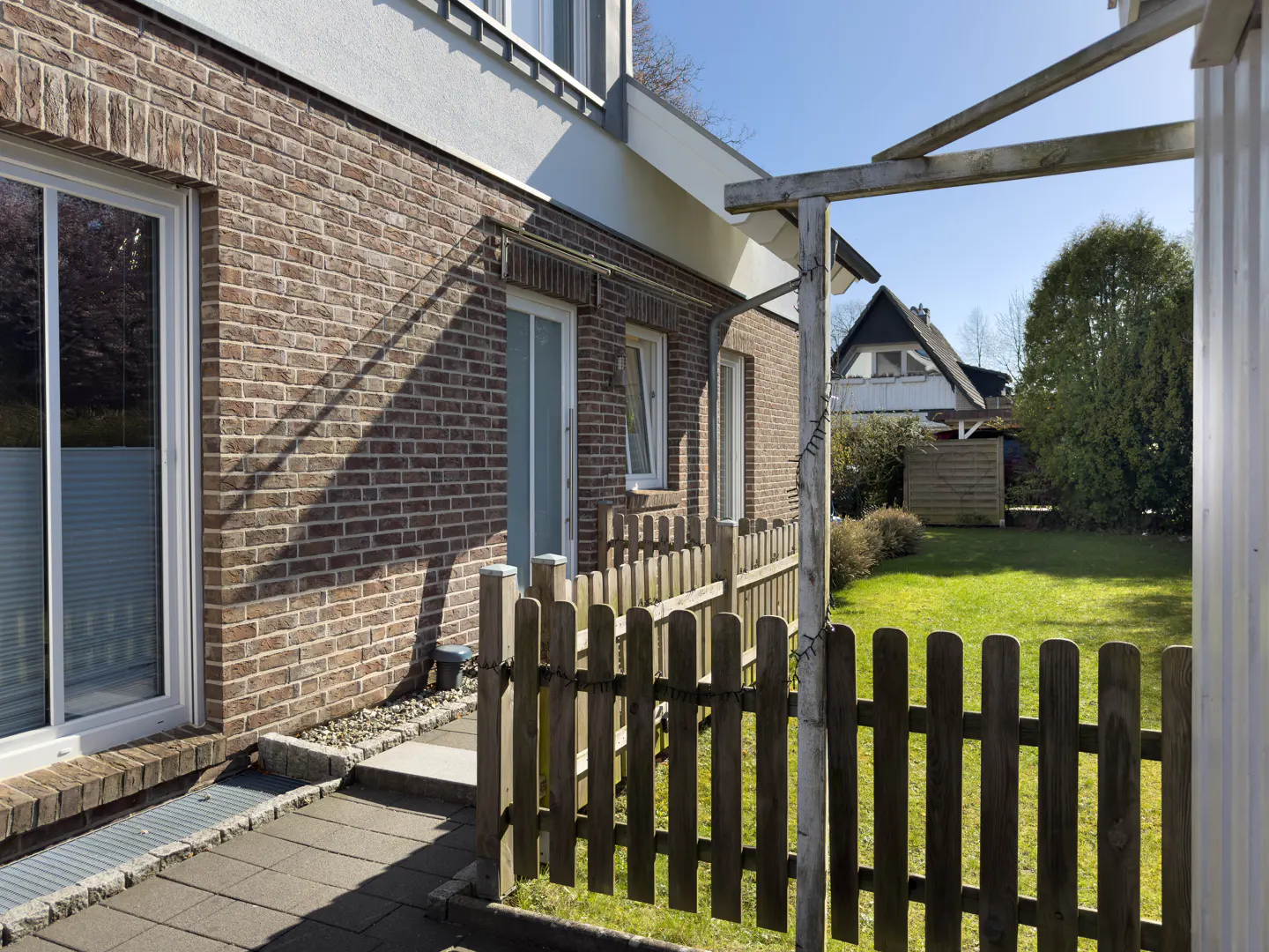 Exterior view of a brick house with a wooden fence and green lawn under a blue sky.