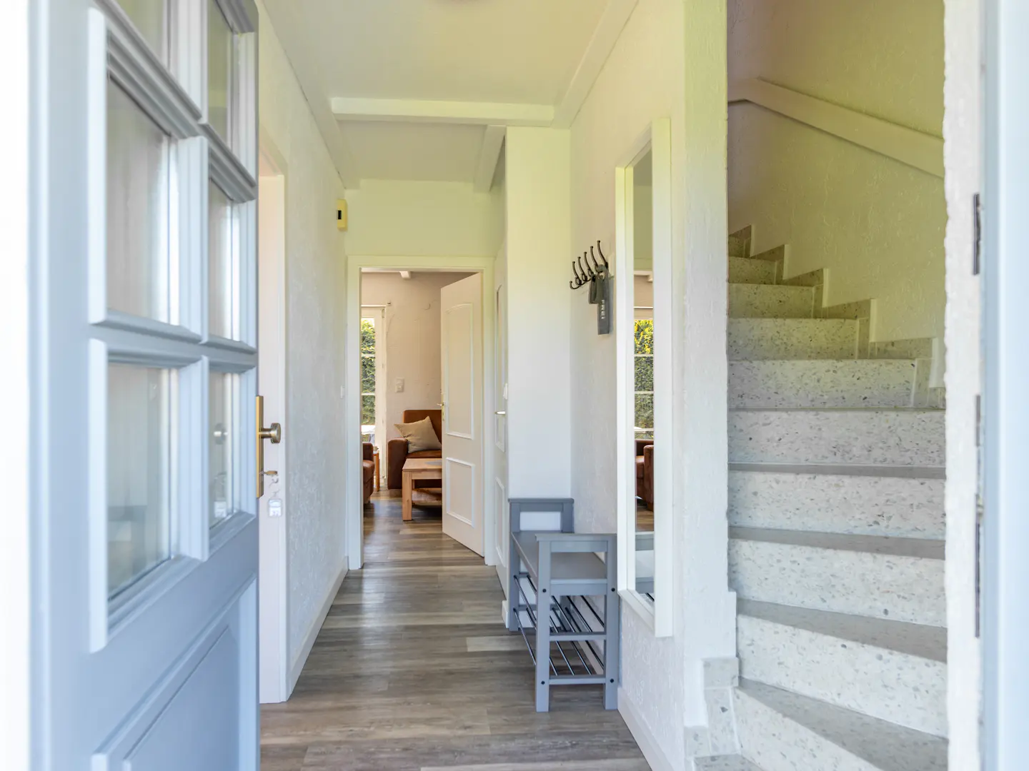 Open front door view of a bright hallway with stairs, a bench, and a glimpse into a living room.