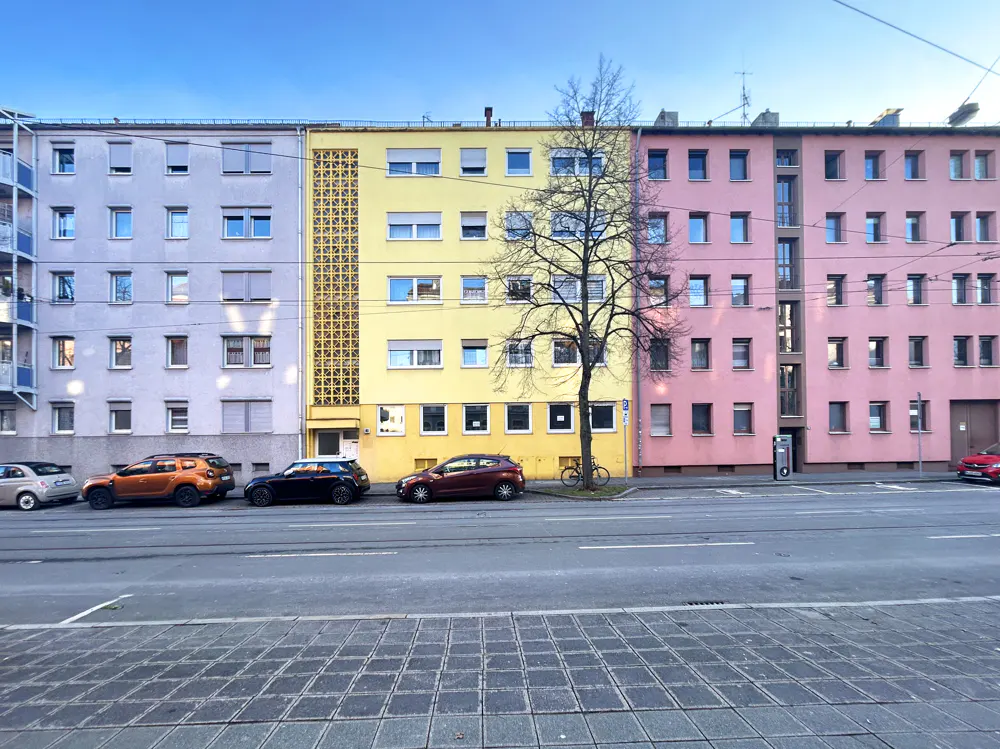 Row of colorful buildings in lavender, yellow, and pink, with parked cars and a bare tree along the street.