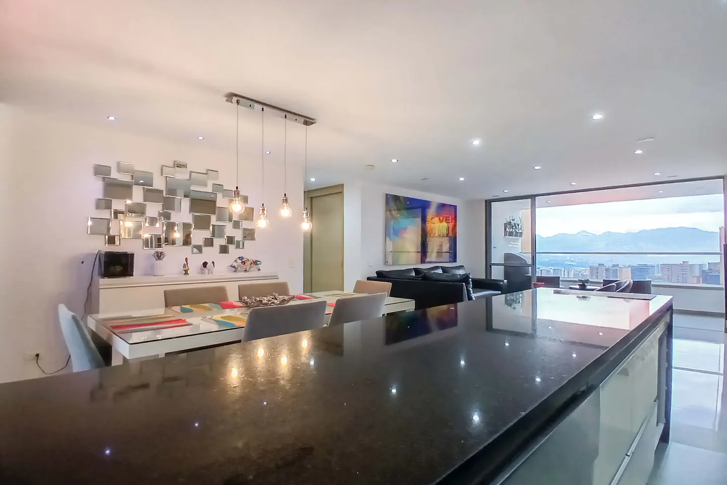 Open-concept living space with a dining table, black kitchen island, and city view. A mirror art piece hangs above a white sideboard.