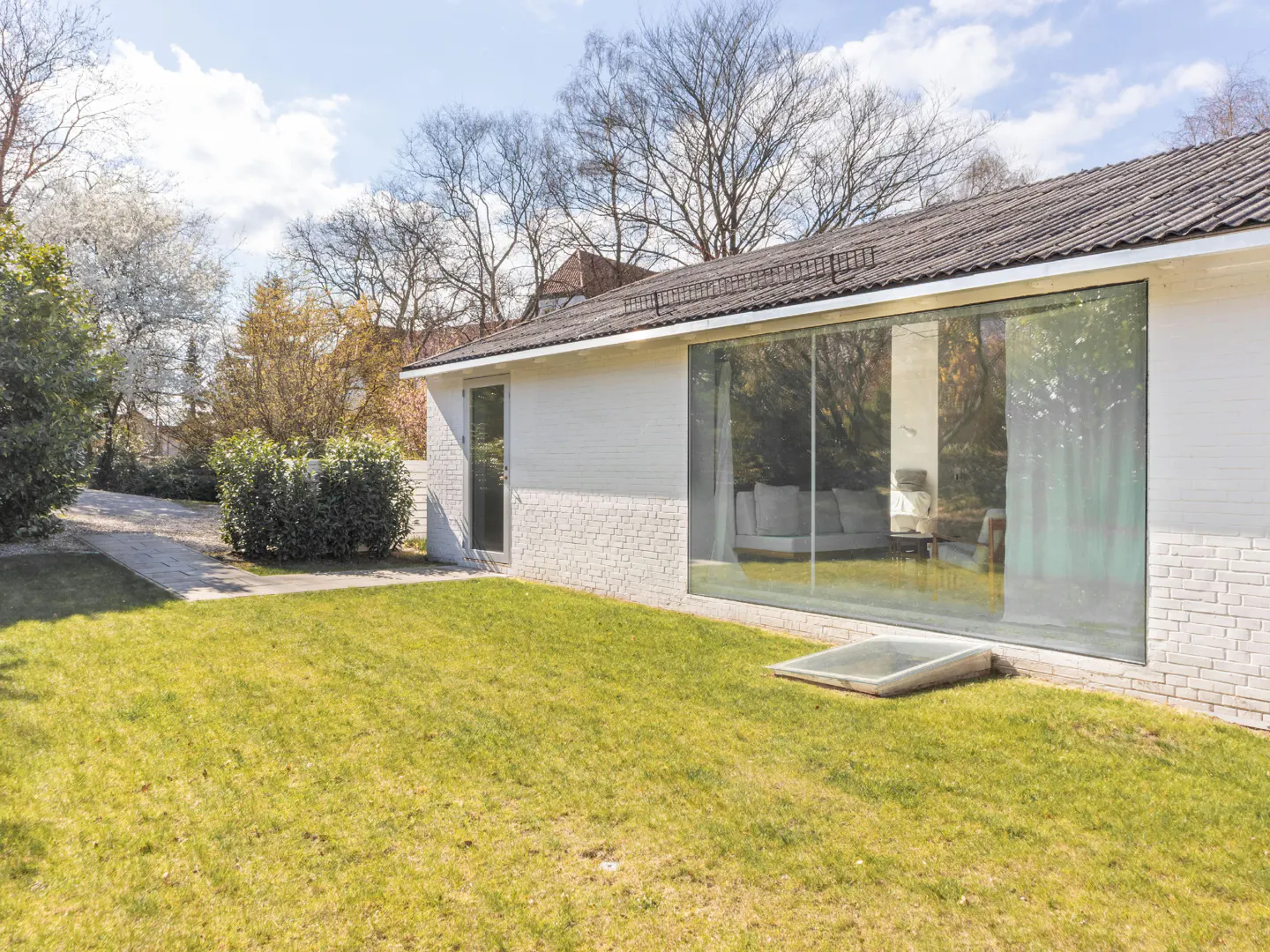 Exterior view of a modern white brick house with a large glass window, green lawn, and trees.