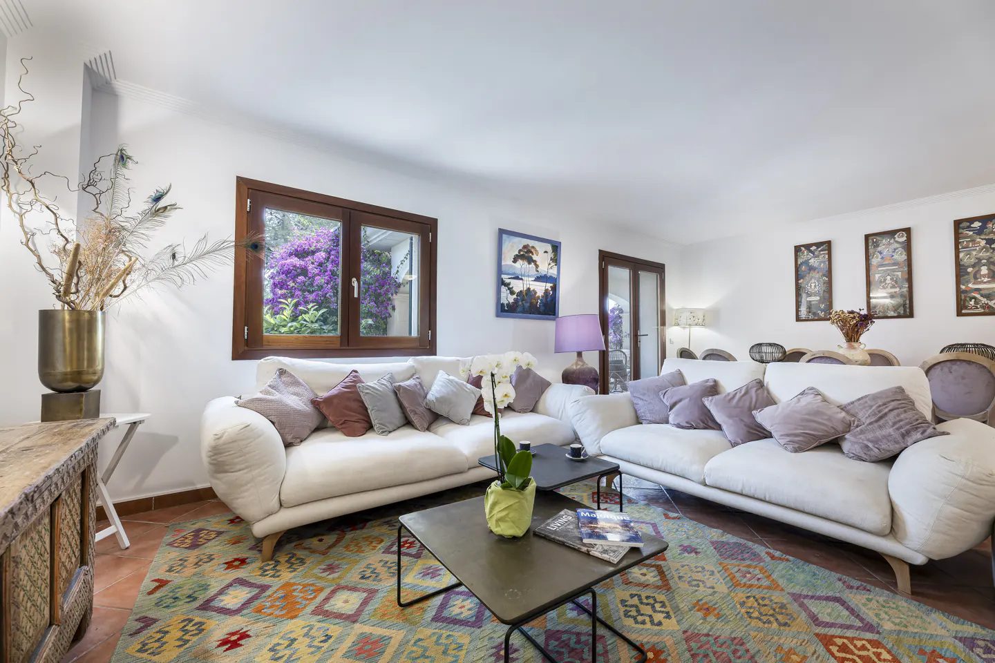 Bright living room with two white sofas, colorful rug, and dark wood window framing purple flowers.