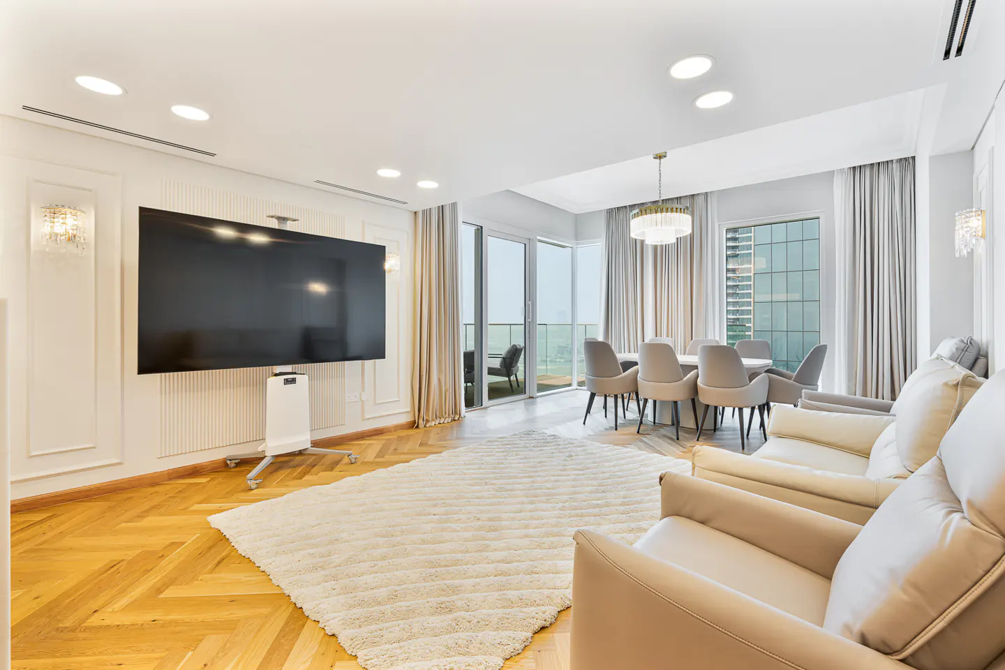 Bright, modern living room with a large TV, dining table, and beige sofas. A white rug covers the herringbone wood floor. Curtains frame a balcony view.
