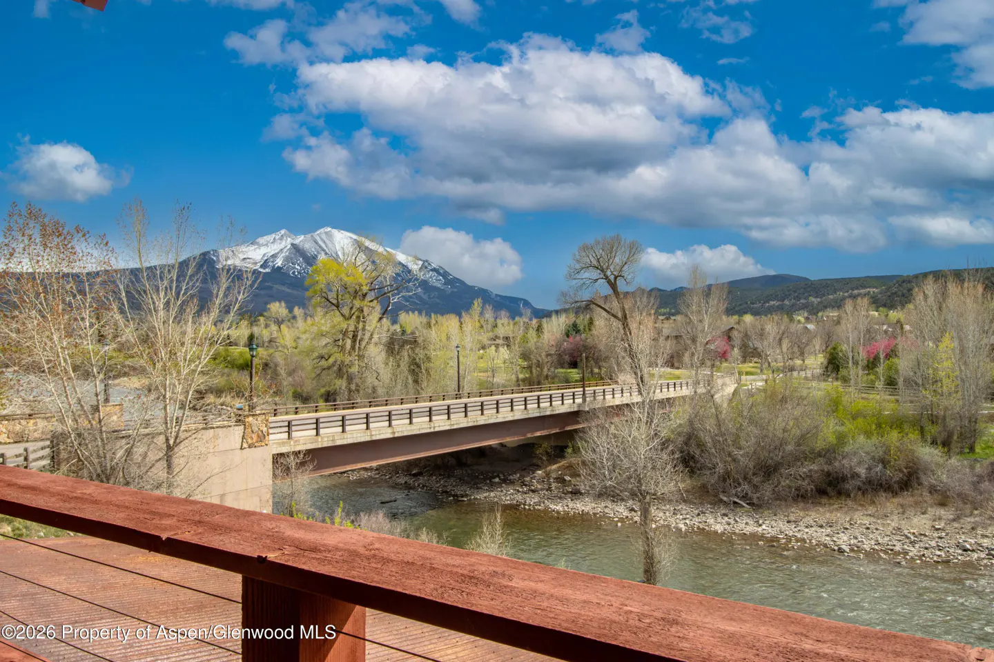 Scenic view from a deck overlooking a river, bridge, and snow-capped mountain under a blue sky with scattered clouds.
