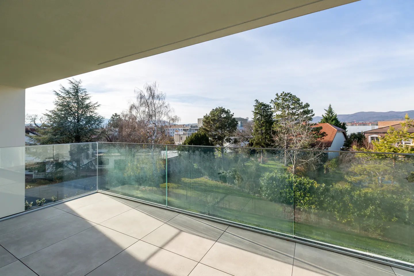Balcony view with glass railing overlooking a green landscape with trees and houses under a blue sky.