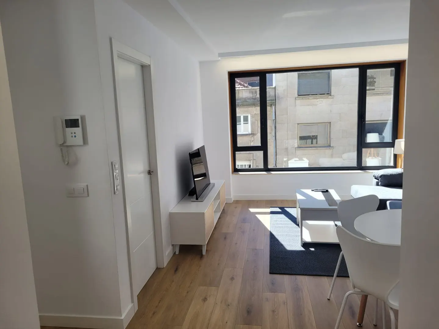 Bright living room with wood floors, white walls, and a large window. A TV sits on a white cabinet. A white table and chairs are in the foreground.