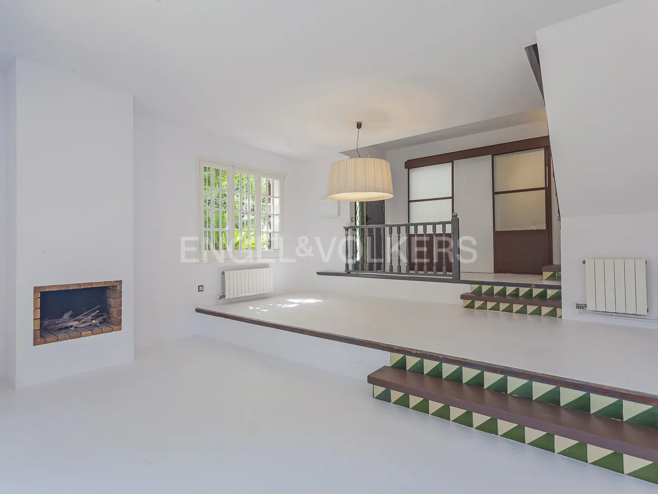Bright, white living room with a fireplace, window, and a raised platform with stairs and a railing. A large lampshade hangs from the ceiling.