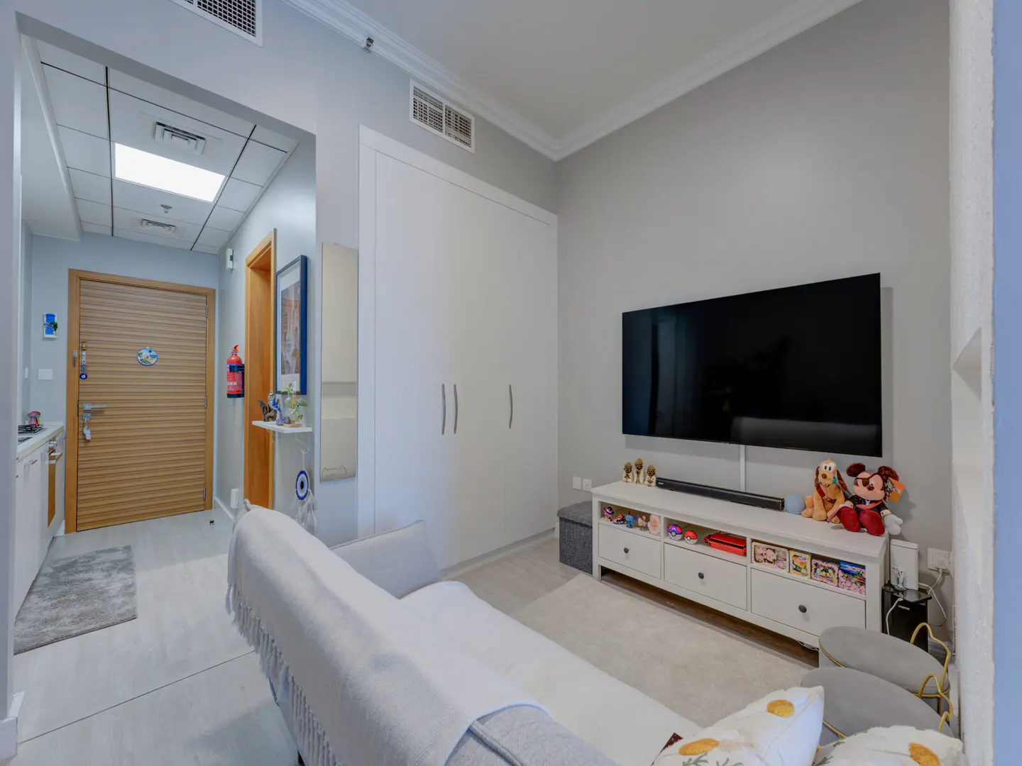 Living room with a gray sofa, white cabinets, and a large TV. Toys are displayed on the white TV stand. A wooden door is visible in the background.