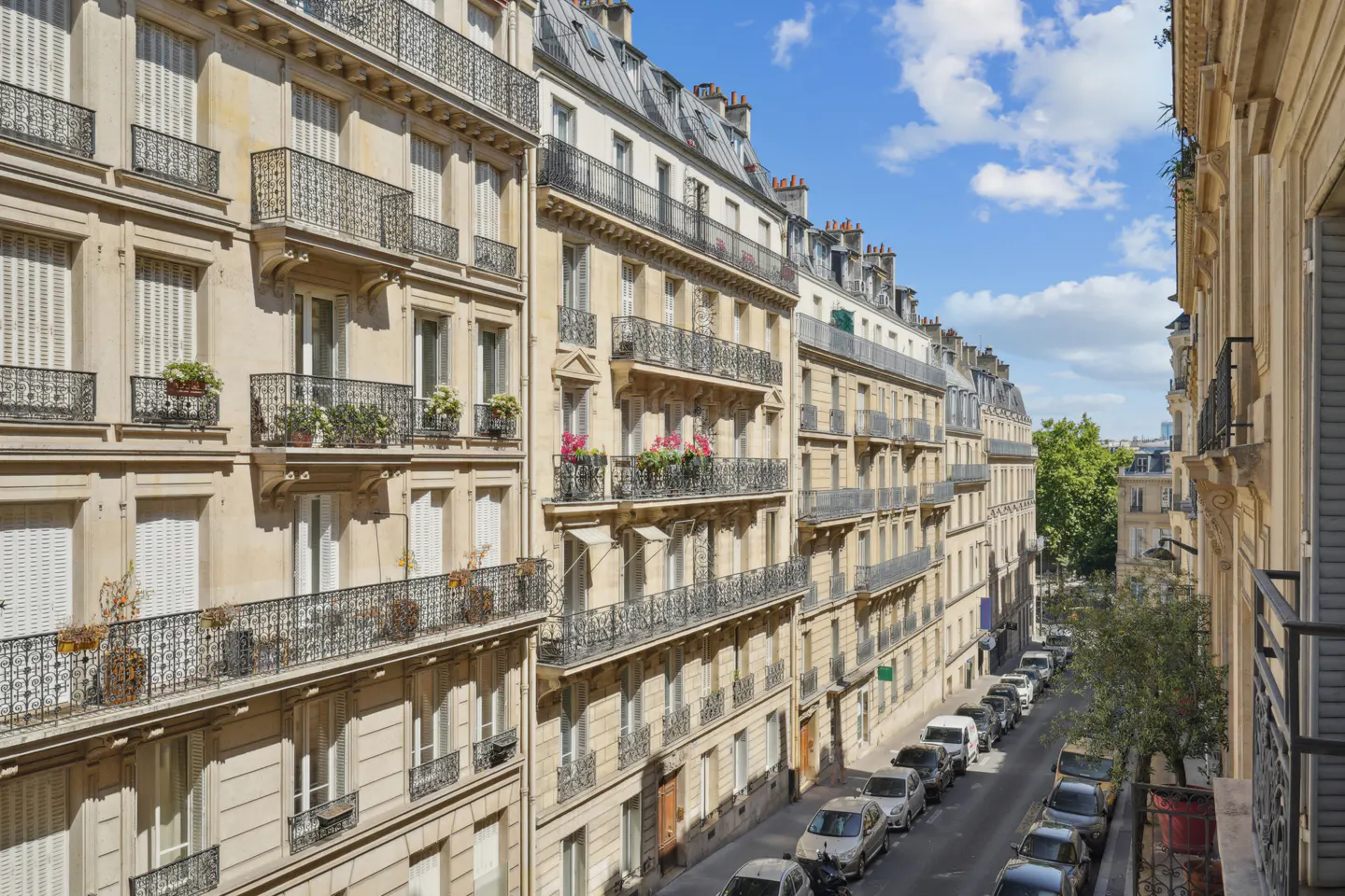 Parisian street view with beige buildings, black wrought-iron balconies, and cars parked along the street under a blue sky.