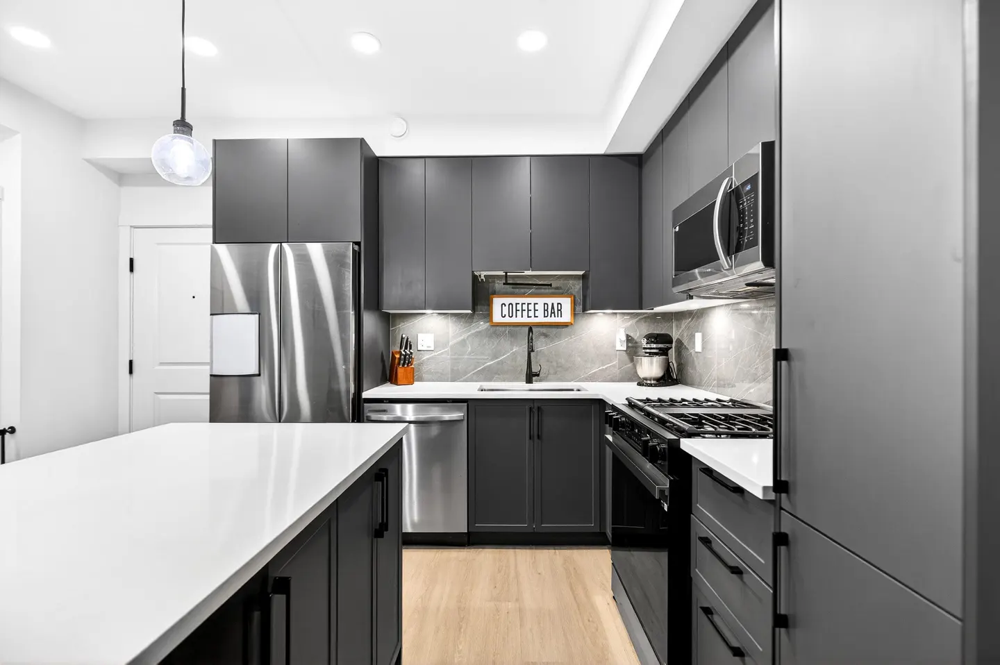 Modern kitchen with gray cabinets, stainless steel appliances, and a white countertop island. A "Coffee Bar" sign hangs above the sink.