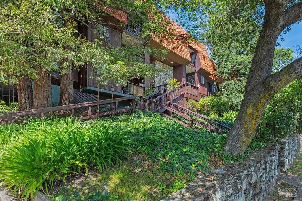 Exterior view of a brown, multi-story building with a wood-shingled roof, surrounded by lush greenery and a stone wall.