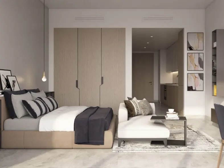 Studio apartment with a bed, white chaise lounge, and kitchenette. Neutral colors and modern decor create a cozy, inviting space.