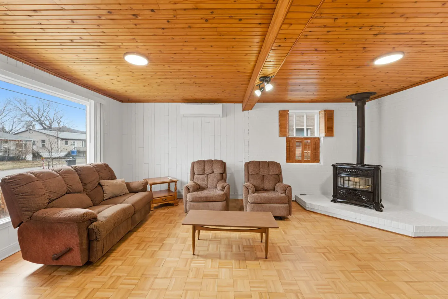 Living room with wood-paneled ceiling, brown sofa and chairs, wood floors, and a black fireplace. A window shows a view of houses outside.