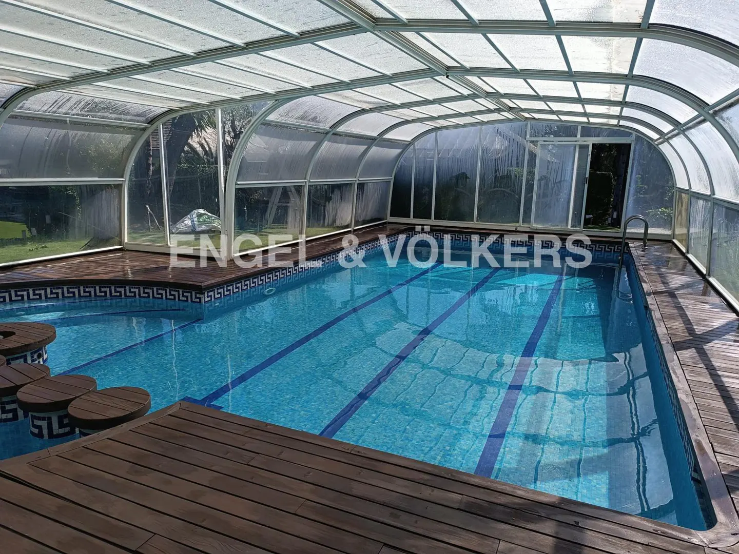 Indoor swimming pool with blue water, wooden deck, swim-up bar stools, and a retractable arched glass enclosure.