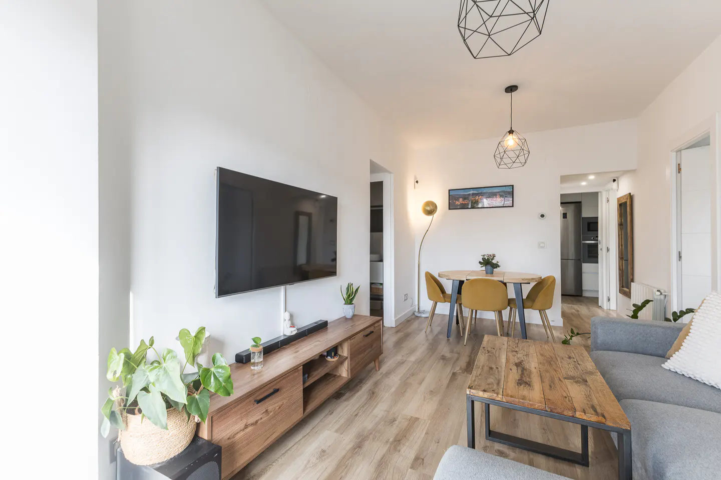 Bright living room with wood floors, white walls, and modern furniture. A large TV hangs above a wooden console. A dining table and gray sofa are visible.