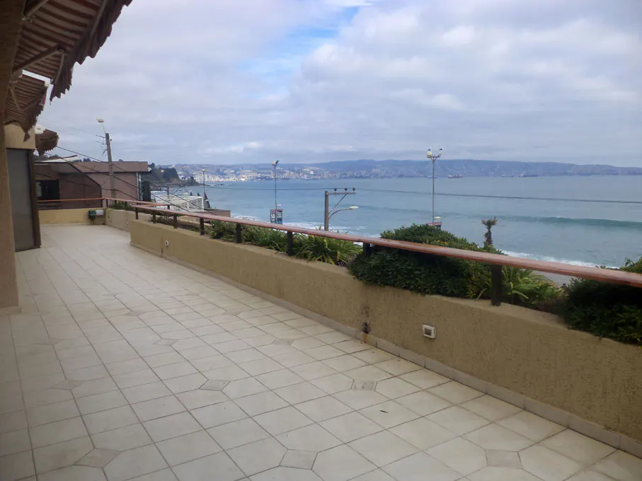Ocean view from a tiled balcony with a low wall and wooden railing. City skyline visible across the water under a cloudy sky.