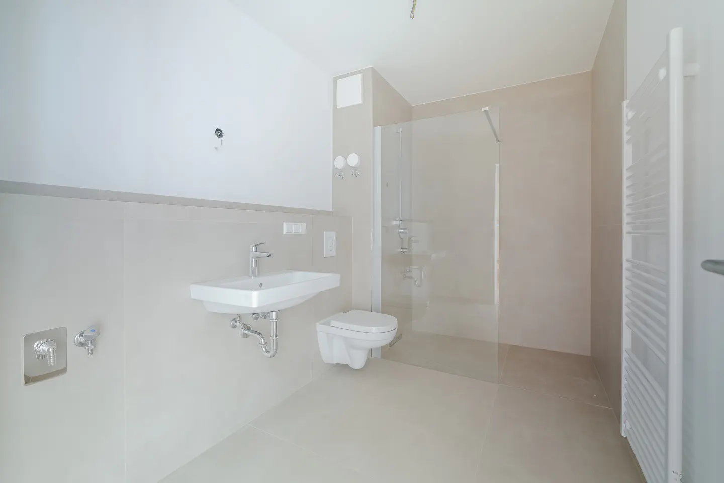Bright, modern bathroom with white sink, toilet, and shower. Beige walls and floor tiles. A white towel rack is on the right.