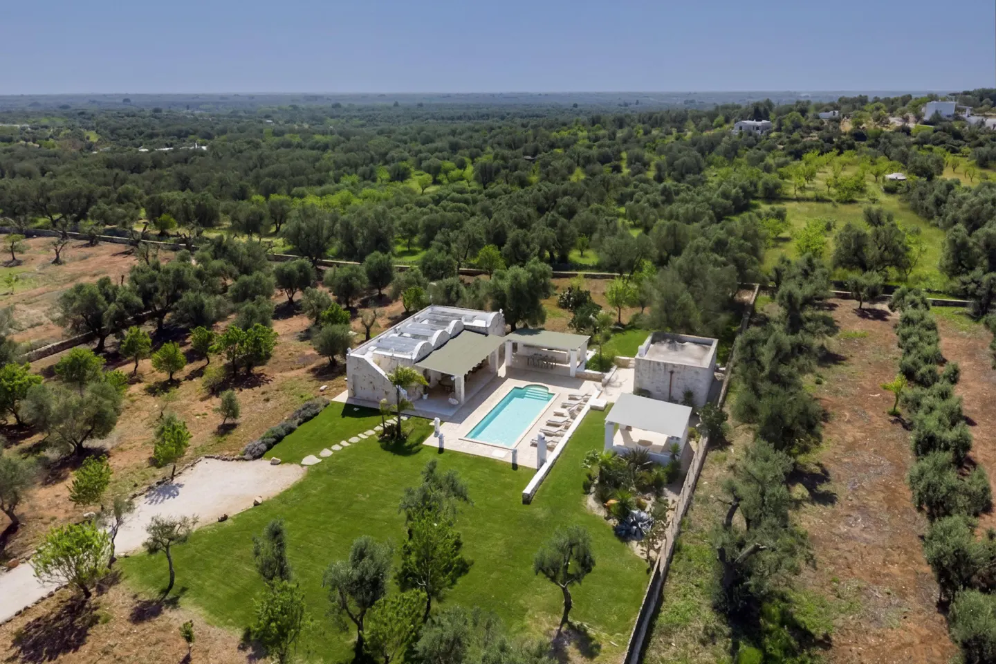 Aerial view of a white villa with a pool surrounded by green lawns and olive trees under a blue sky.