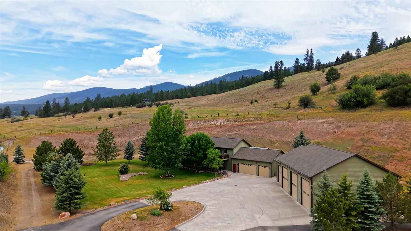 A green house with a large garage sits on a grassy hill under a blue, cloudy sky. Mountains are in the background.