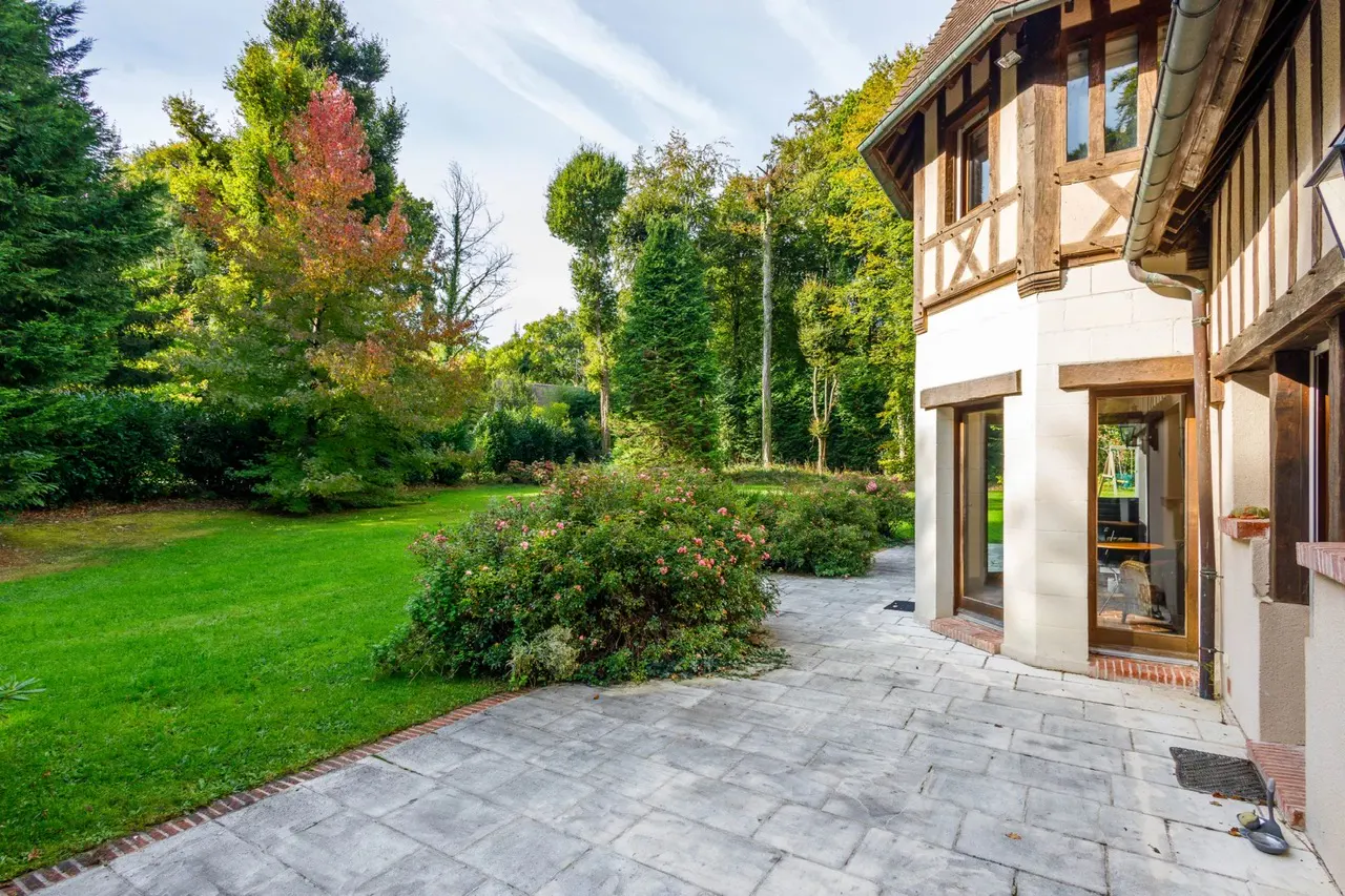 Exterior view of a Tudor-style home with a stone patio, green lawn, and trees in the background.