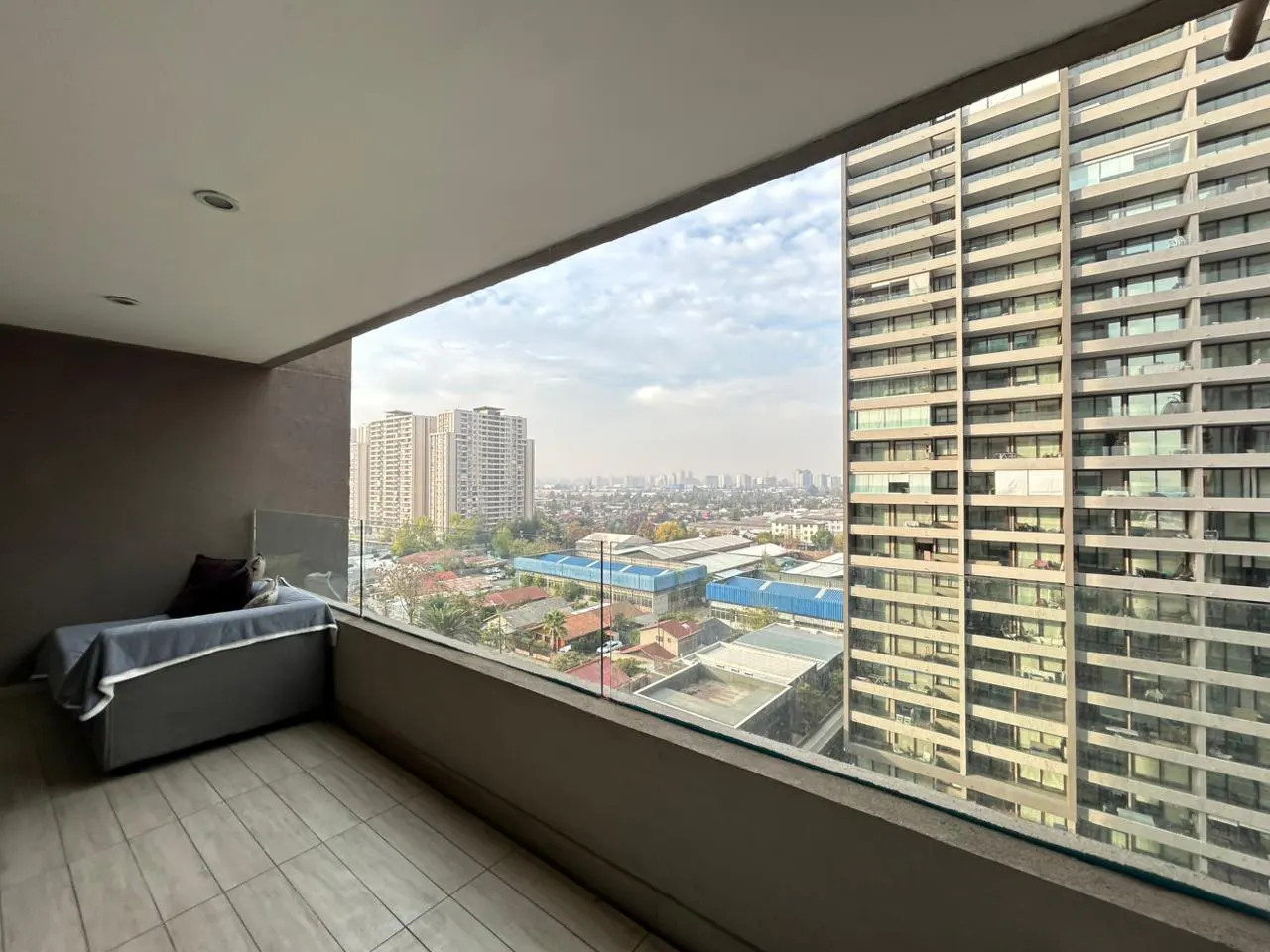 Balcony view of a city skyline. Gray daybed with a pillow and blanket. Tall buildings in the background.