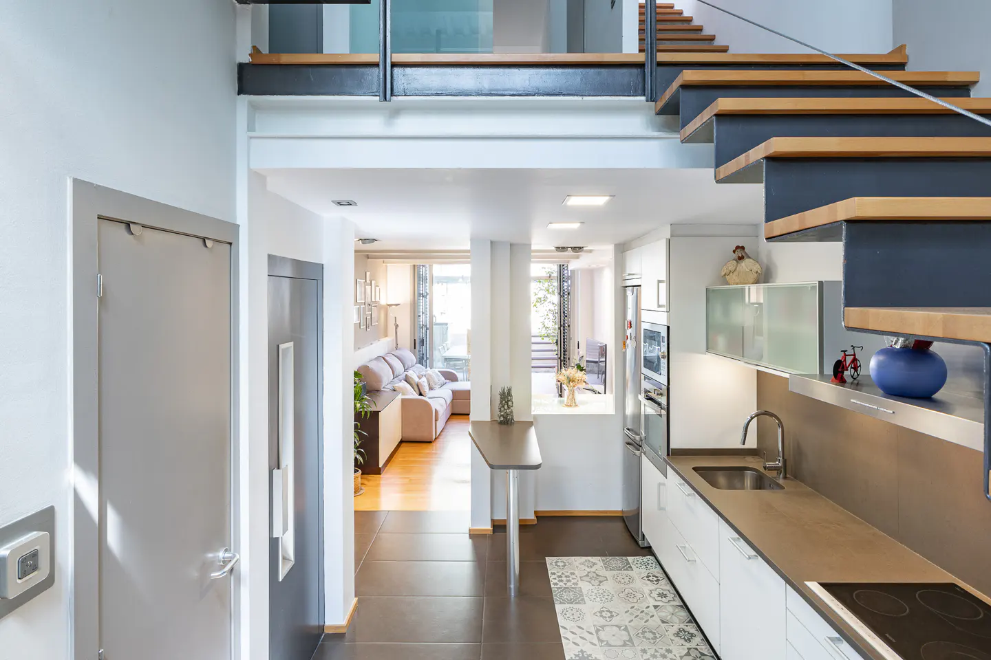 Interior view of a modern home with stairs, kitchen, and living room. White walls, stainless steel appliances, and brown tile flooring.