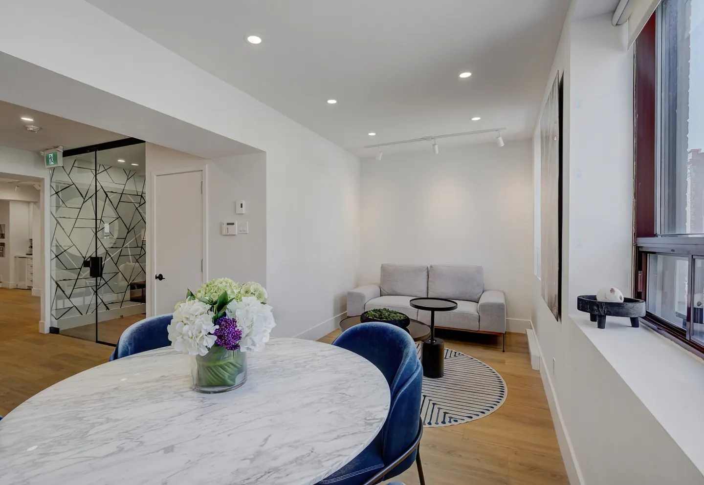 Bright, modern living space with a marble table, blue chairs, and a gray sofa. A glass-walled office is visible in the background.