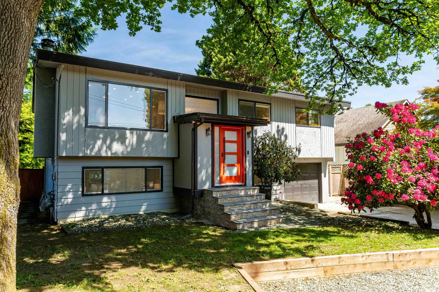 Two-story gray house with a bright orange front door and stone steps leading up to it. Green lawn and trees surround the house.
