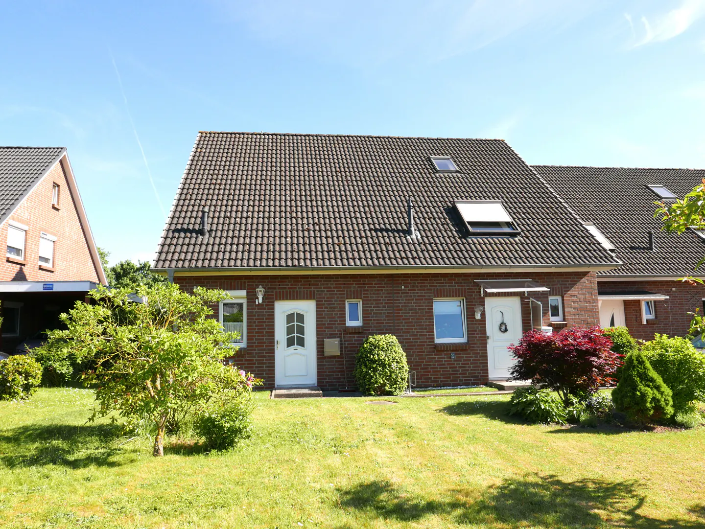 A two-story red brick duplex with a dark tiled roof, white doors, and a green lawn under a clear blue sky.