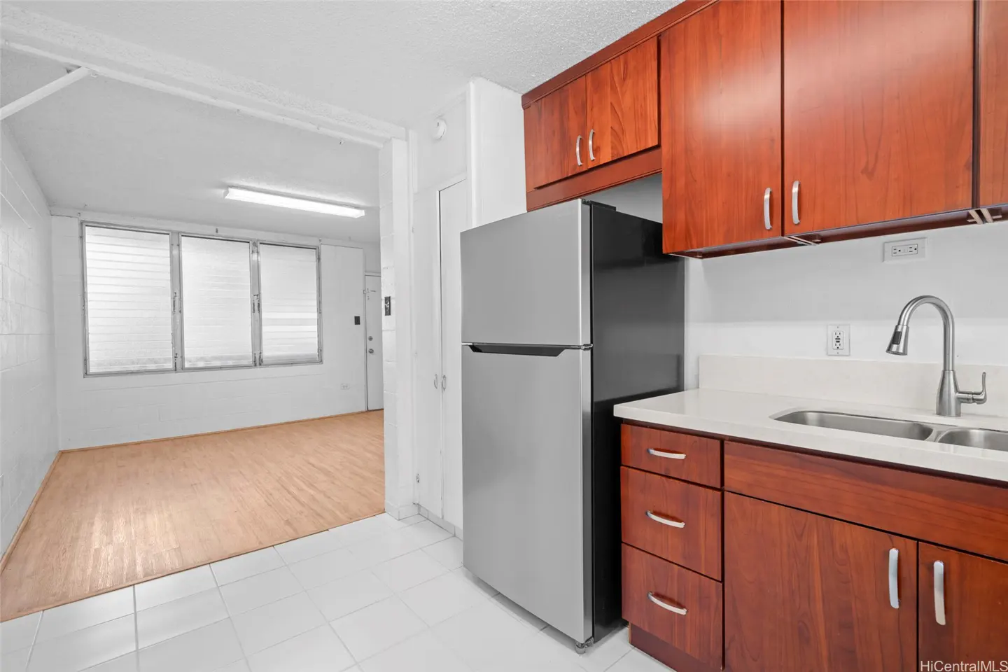 A kitchen with cherry wood cabinets, a stainless steel refrigerator, and a white countertop with a sink. A doorway leads to a bright, empty room.