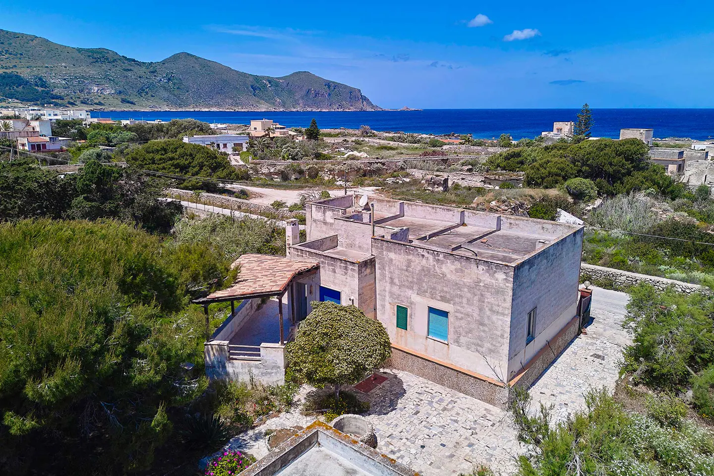 Aerial view of a beige house with a flat roof and tiled porch, surrounded by greenery near a blue sea and mountains.
