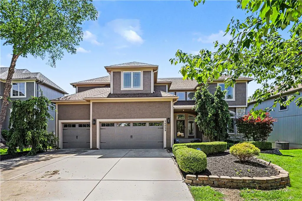 Two-story stucco house with a three-car garage and manicured landscaping on a sunny day.