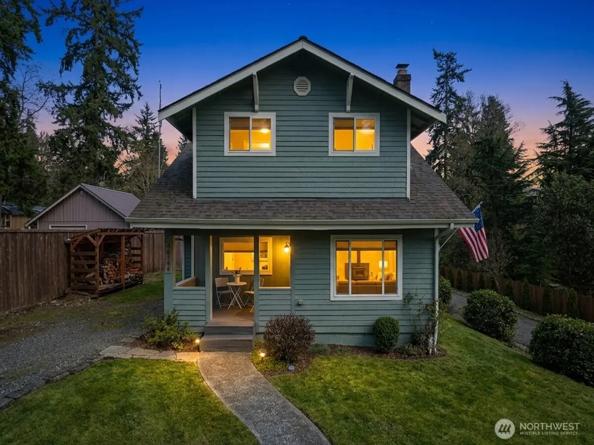 Two-story teal house with a gray roof and white trim, lit windows, and an American flag at dusk.
