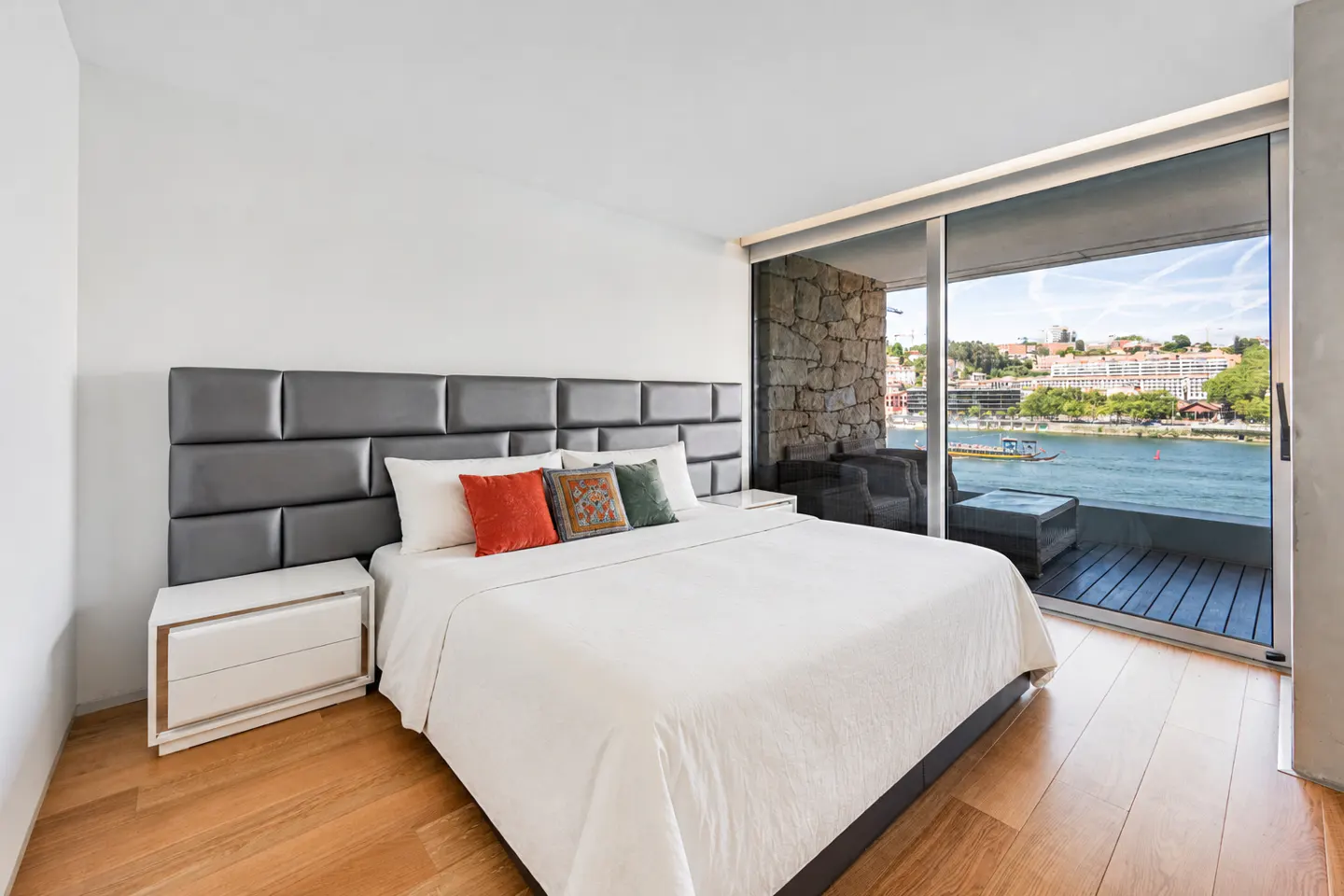 Modern bedroom with a white bed, grey headboard, and floor-to-ceiling windows overlooking a river and city skyline.