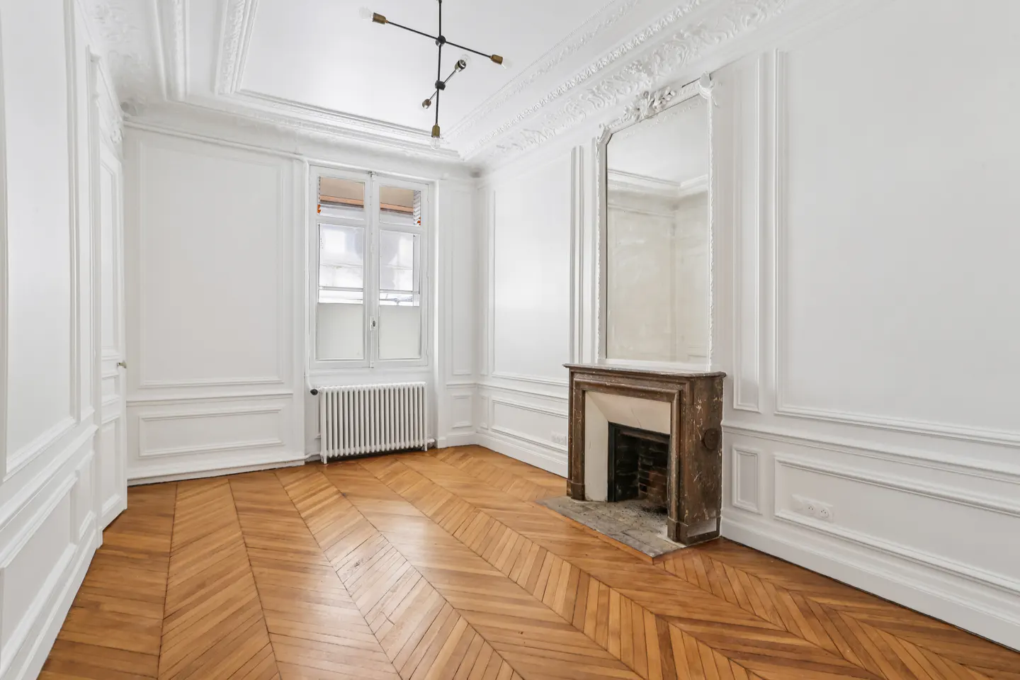 Bright room with white paneled walls, herringbone wood floors, a marble fireplace, tall mirror, and modern light fixture.