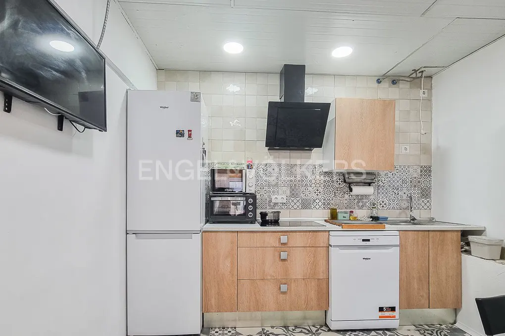 A kitchen with white walls, light wood cabinets, and a black range hood. A white refrigerator stands next to a microwave and oven. A TV is mounted on the wall.