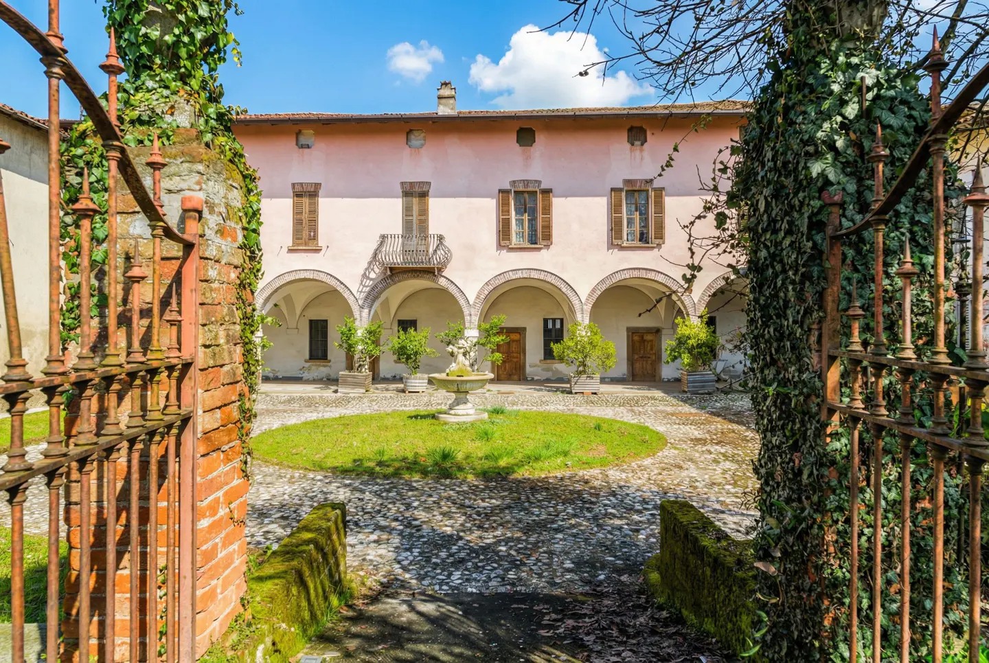 Courtyard view through an iron gate. A pink building with arched openings surrounds a green lawn with a fountain.